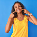 Brunette woman in a yellow tanktop smiles and points to her teeth after going to an affordable dentist in Mililani, HI