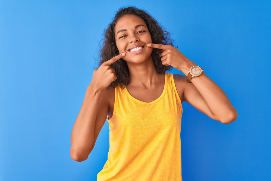 Brunette woman in a yellow tanktop smiles and points to her teeth after going to an affordable dentist in Mililani, HI