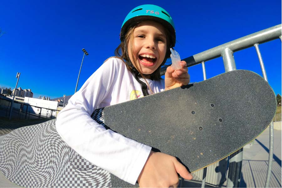 young girl wearing a helmet, holding a skateboard under her arm, holds up her mouthguard and grins a